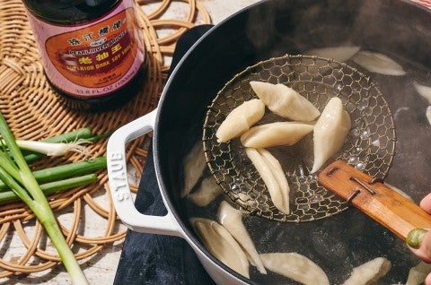 A handheld noodle mesh scoop picking up hand cut noodles from a pot of boiling water seen from above the food. - select to zoom