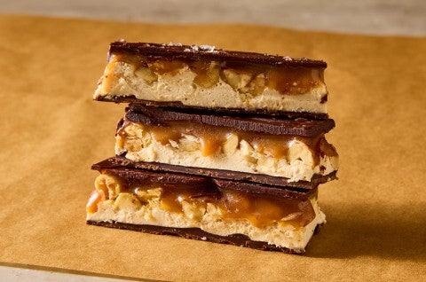 Three sliced chocolate peanut nougat bars stacked on brown parchment and showing the layers of chocolate, nougat, caramel, and peanuts. - select to zoom
