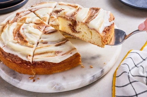 Removing a slice of Cinnamon Roll Cake from the cake showing the cinnamon sugar layer inside the golden cake and the icing on top. - select to zoom