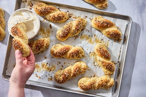 A hand dipping an Everything Bagel Twist into cream cheese along with other bagel twists spread out on a sheet tray and parchment paper. - select to zoom