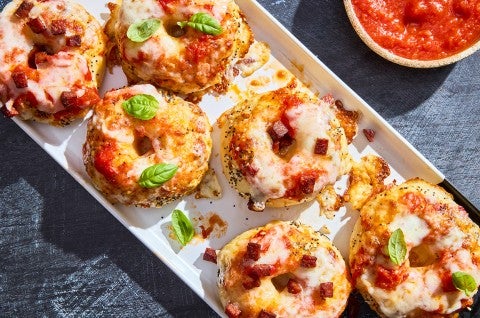 Delicious mini pizza bagels arranged on a plate showing melty cheese, pepperoni, and basil with a bowl of marinara in the top of the photo. - select to zoom