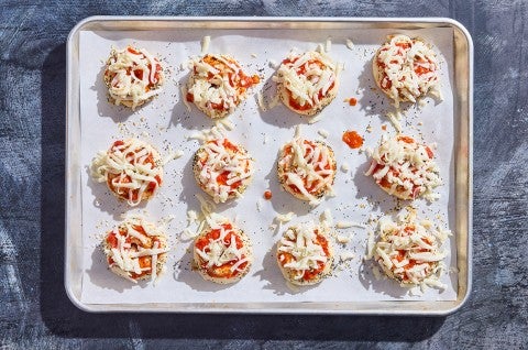 12 uncooked prepared mini pizza bagels on a sheet tray ready to go into the oven. - select to zoom