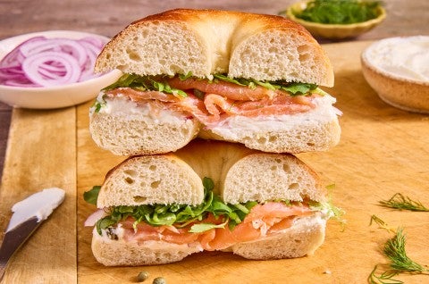A bagel sandwich sliced in half and stacked with the interior side showing layers of smoked salmon, greens, cream cheese, and a light and chewy bagel interior. - select to zoom