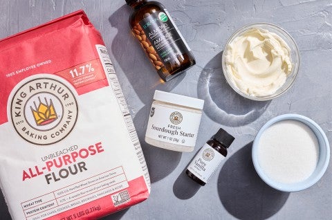 Ingredients to make Sourdough Sugar Cookies - flour, sourdough starter, almond extract, vanilla extract , sugar, and cream cheese - shown on a counter from above. - select to zoom