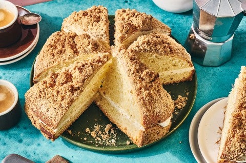 A round, Cream-Filled Coffee Cake, sliced on a dark plate with other breakfast elements spread around a table. - select to zoom