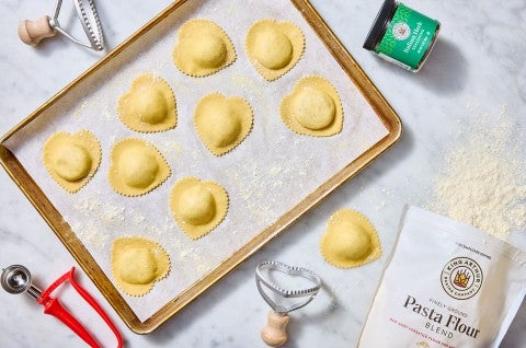 Homemade Cheese Ravioli shown cut into heart shapes and organized on a sheet tray covered in parchment with ingredients and tools strewn about a light marble counter. - select to zoom