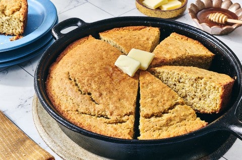 Sourdough Cornbread in a cast iron skillet on a table with a small slice seen in the corner on a blue plate. - select to zoom