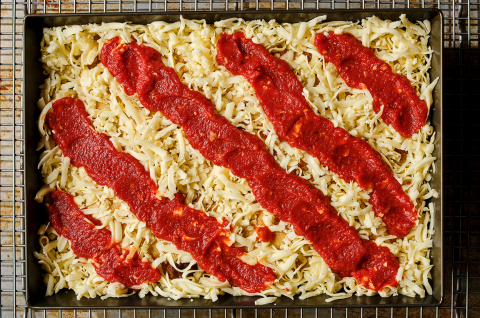 Preparing Detroit style pizza in a classic dark pizza pan with strips of sauce over the top of cheese. - select to zoom