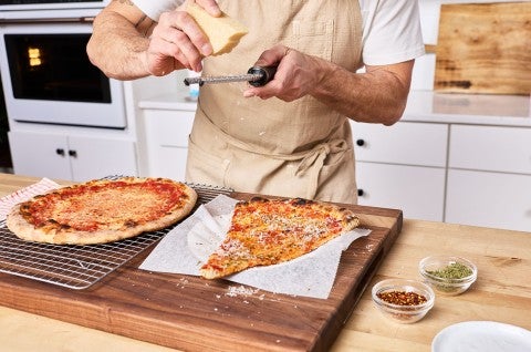 Martin grating cheese over a slice of New York-Style Pizza on a counter. - select to zoom