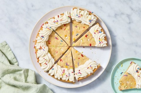 Sliced Confetti Sugar Cookie Cake on a marble countertop with a piece on a plate. - select to zoom