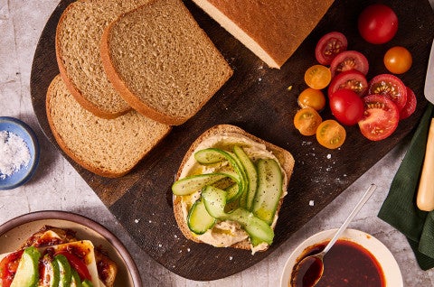 Classic 100% Whole Wheat Bread sliced on a cutting board with tomatoes, cucumbers, and other elements. - select to zoom