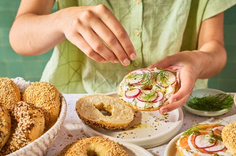 Someone sprinkling seasoning on a dressed bagel. - select to zoom
