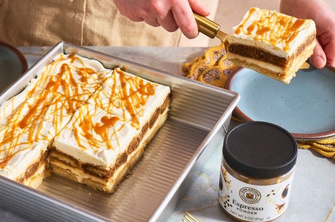 Putting a slice of Caramel Macchiato Tiramisu on a plate with the rest of the baked good in a baking sheet and a jar of King Arthur Baking Espresso Powder in the foreground. - select to zoom