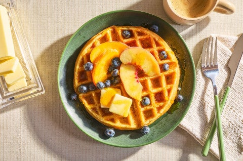 A Classic Buttermilk Waffle on a green plate and topped with butter, syrup, peaches, and blueberries. - select to zoom