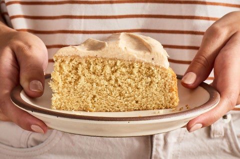 A slice of Frosted Brown Butter Cake being held on a plate seen from the side - select to zoom