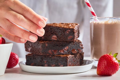 Fudge Brownies stacked on a plate with someone picking up the top one and strawberries in the foreground. - select to zoom