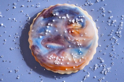 A Northern Lights Iced Sugar Cookie on a blue background surrounded by sprinkles of sparkling sugar - select to zoom