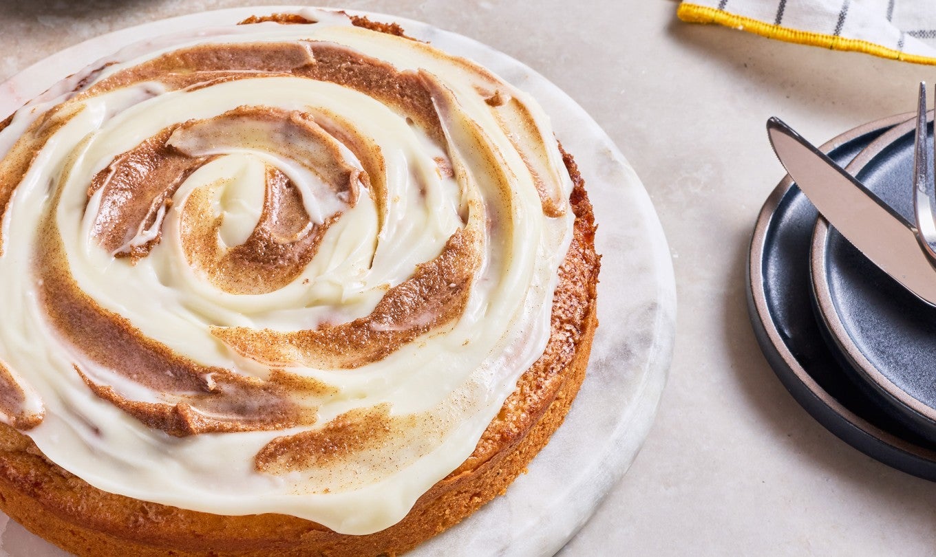 A cinnamon roll cake on white marble round cake platter with a knife and fork atop a plate next to it.