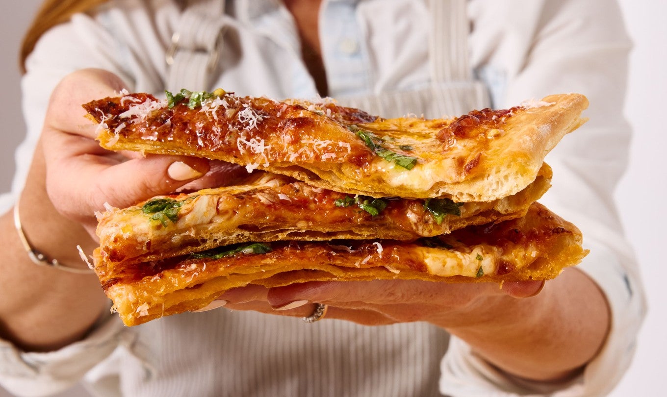A person holding three slices of flaky puff crust pizza and showing the laminated and crispy crust, and the melty cheese.