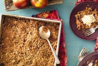 A pan of apple crisp on a table with a few apples and two plates of apple crisp with ice cream