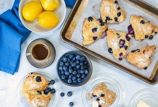 Fresh blueberry scones on a baking sheet next to a bowl of blueberries, a few lemons, a coffee, and butter.