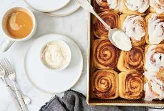 A tray of cinnamon buns with a bowl of frosting next to a cup of coffee