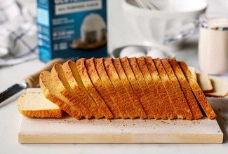 A loaf of gluten-free sandwich bread, sliced on a bread board with a box of gluten-free flour in the background