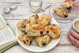 A plate of cream scones next to a book and a person holding a mug of coffee