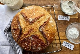 A loaf of sourdough bread on a cooling rack next to a starter and a bowl of baking powder and baking soda