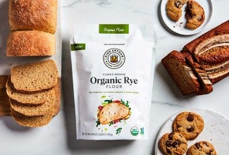 A loaf of rye sandwich bread, rye banana bread, and rye chocolate chip cookies next to a bag of rye flour