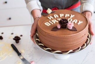 A baker presenting a birthday cake topped with modeling chocolate letters and flowers