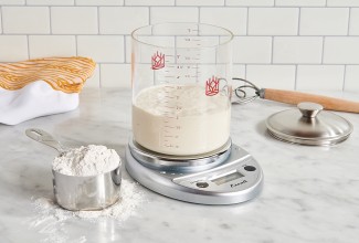 Glass sourdough crock on digital scale next to measuring cup full of flour