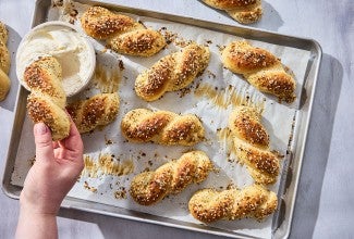 A hand dipping an Everything Bagel Twist into cream cheese along with other bagel twists spread out on a sheet tray and parchment paper.