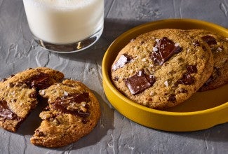 Beautifully baked High-Altitude Chocolate Chip Cookies shown on a plate and counter with a glass of milk in the background.