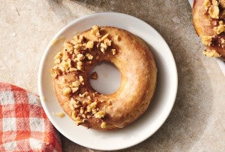 A Maple Donut on a plate with half coated in broke nut meat and the other half glazed.