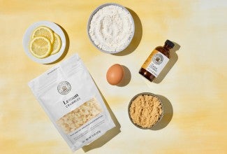 Assorted ingredients for Chewy Lemon Blondies on a yellow surface, including lemon extract, slices of lemon, lemon crumbles, an egg, and two types of sugar in small bowls