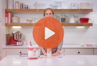 A person standing behind a counter with sourdough starter ingredients and a play icon over the image. - select to zoom