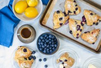 Fresh blueberry scones on a baking sheet next to a bowl of blueberries, a few lemons, a coffee, and butter.