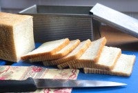 Loaf of white sandwich bread, sliced, on a cutting board in front of a pain de mie pan.