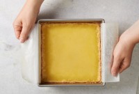 Hands using parchment paper sling to remove lemon bars from square pan