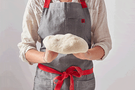 A baker holding a piece of dough draped over the back of their knuckles, slowly turning the dough to stretch it into a round. 