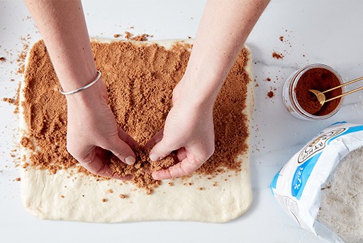 A baker sprinkling a mixture of cinnamon and brown sugar over a rectangle of dough. 