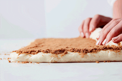 A low side-angel shot of baker rolling the filling-topped dough into a log, showing the loose spiral of dough and filling. 