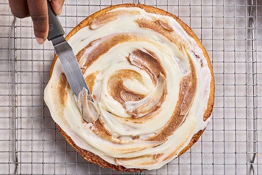 Cinnamon Roll Cake on a wire rack with a person spreading the frosting on top.