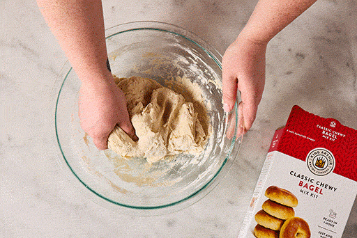 Overhead view of shaggy bagel dough being mixed in a bowl with a box of King Arthur Baking Chewy Bagel Mix in the bottom right corner of the frame.