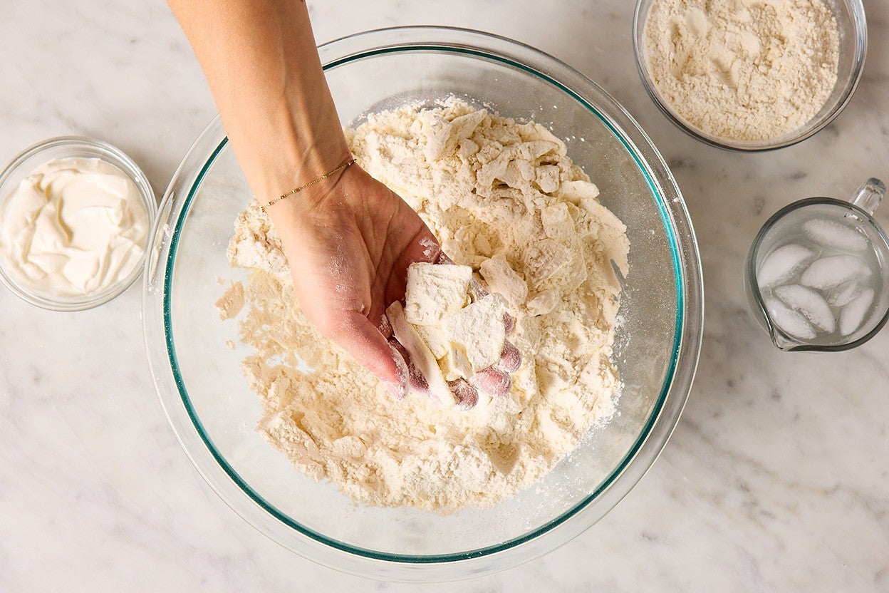 A hand holding bits of cold butter within a dough mix to highlight the thing shaved pieces of butter in a flaky biscuit recipe.