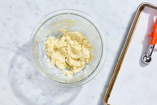 Gulab Jamun dough in a clear bowl with a small scoop and parchment on a baking tray.
