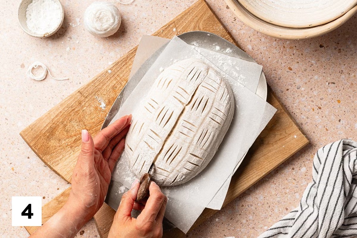 Sourdough loaf with a basket weave design scored into it, with a baker scoring a long line down the center