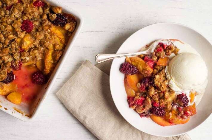 A pan of freshly baked, crumb-topped Summer Fruit Crisp next to a bowl of fruit crisp with a spoon