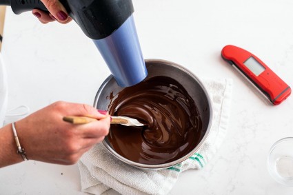 A baker using a hair dryer to warm chocolate and keep it in temper